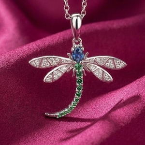 May include: A silver dragonfly pendant necklace. The dragonfly features a blue gemstone head, green gemstone body, and wings adorned with clear gemstones. The necklace is displayed on a deep pink fabric.
