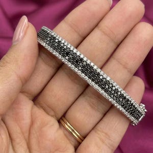 May include: A silver bracelet with a row of black stones in the center, bordered by rows of white stones. The bracelet is held against a pink background.