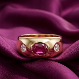 May include: A wide gold band ring featuring a central oval-cut vibrant pink gemstone set horizontally. The ring is accented by two smaller round-cut clear white diamonds, one positioned on each side of the central pink stone in a bezel setting.