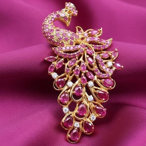 Könnte beinhalten: Goldfarbene Pfauenbrosche mit detailliertem Design. Der Körper und die Schwanzfedern des Pfaus sind mit rosa Edelsteinen und kleinen weißen Akzenten verziert. Die Brosche ist auf tiefrosa Stoff gesetzt.