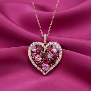 May include: A gold-coloured heart pendant necklace with a chain. The heart is filled with pink and red heart-shaped gemstones, surrounded by a halo of small, clear stones. The necklace rests on a pink satin fabric.