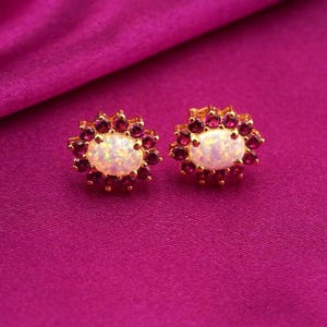 Puede incluir: Un par de pendientes de botón dorados. Cada pendiente presenta una piedra central ovalada y opalescente rodeada de pequeñas gemas redondas de color rojo intenso. Los pendientes están sobre un fondo magenta vibrante.