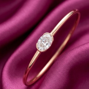 May include: A rose gold bangle bracelet featuring a large, oval-cut diamond. The diamond is set in a bezel setting and is the focal point of the bracelet. The bracelet is displayed on a deep purple satin surface.