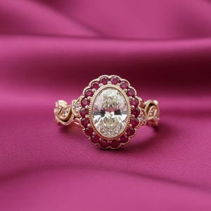 May include: A gold ring with an oval-cut, clear gemstone center, surrounded by small, round, red gemstones. The ring has intricate detailing on the band and a pink background.