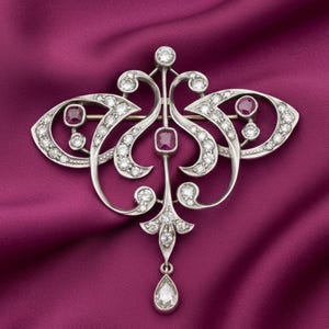 May include: A detailed silver brooch with a symmetrical design, set with numerous small diamonds and several red gemstones. A teardrop-shaped diamond hangs from the bottom. The brooch is displayed against a deep purple fabric, creating a striking contrast.