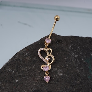 Op de afbeelding: Goudkleurige navelring met hartvormige bedels. De bedels zijn versierd met kleine, roze edelstenen en heldere kristallen. De boven- en onderkant van de ring hebben een kleine, ronde gouden bal.