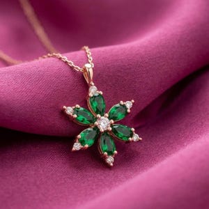 May include: A gold-toned necklace with a flower pendant. The pendant features emerald-green gemstones arranged in a floral pattern, with a central white gemstone and smaller white gemstones accenting each petal. The necklace rests on a pink fabric.