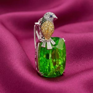 May include: A detailed jewelry piece featuring a bird perched on a large, rectangular green gemstone. The bird has a silver, gold, and blue color scheme. The gemstone is set in a silver metal frame, all against a magenta background.