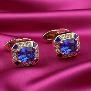 May include: A pair of gold-toned cufflinks with a square, blue gemstone center. The cufflinks have a gold border with small, clear stones and dark blue accents. The background is a vibrant pink fabric.