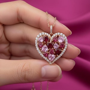 May include: A gold necklace featuring a heart-shaped pendant. The pendant is adorned with pink and red gemstones, encircled by a border of small, clear stones. The necklace is held in a hand against a pink backdrop.