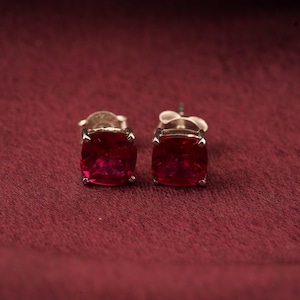 May include: A pair of square-cut, ruby-red gemstone stud earrings. The gemstones are set in a silver-colored metal. The earrings are displayed on a textured, maroon-colored surface.