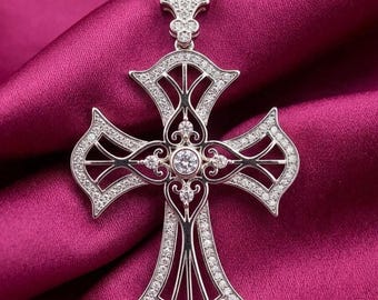 May include: A silver-toned cross pendant necklace with a gold chain. The cross is encrusted with small, clear gemstones and features a large, round gemstone at the center. The cross is set against a deep pink fabric.