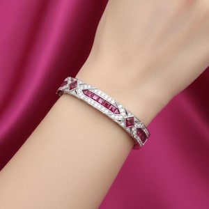 Può includere: Un braccialetto d'argento con un design geometrico con pietre preziose rosse rettangolari e quadrate, intervallate da piccole pietre preziose rotonde e trasparenti. Il braccialetto è indossato su un polso su sfondo rosa.