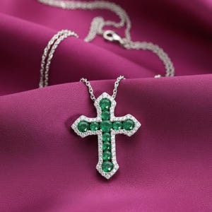 May include: A silver necklace with a cross pendant. The cross is adorned with emerald-green gemstones and surrounded by smaller, clear gemstones. The pendant rests on a deep purple fabric.