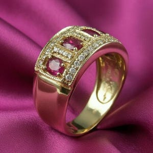 May include: A gold ring featuring three oval rubies and small diamonds. The ring has a wide band and a detailed setting. The gemstones are arranged in a rectangular pattern, with diamonds encircling the rubies. The ring is shown on a pink satin surface.