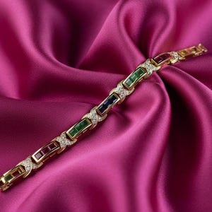 Pode incluir: Pulseira dourada com pedras retangulares alternadas em vermelho, verde e azul, intercaladas com detalhes de diamantes. A pulseira é exibida sobre um tecido de cetim fúcsia vibrante, criando uma estética luxuosa.