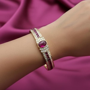 May include: A gold bracelet with a large oval ruby center stone and smaller rectangular rubies and diamonds. The bracelet is on a wrist, showcasing the jewelry's design and color against a pink background.