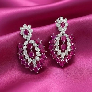 May include: A pair of elegant drop earrings featuring a floral design at the top and a cluster of oval-shaped, deep red gemstones. The earrings are accented with small, clear stones and set against a pink satin background.