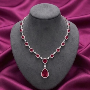 May include: A silver necklace with oval and pear-shaped red gemstones, accented with small white stones. The necklace is displayed on a gray bust against a purple satin background. The pendant features a large, teardrop-shaped ruby.