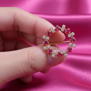 May include: A gold-colored brooch in a circular design, adorned with alternating red gemstones and small, clear crystals. The brooch is held against a pink satin background, showcasing its intricate details.