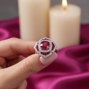 Puede incluir: Anillo de diseño floral con un rubí rojo central, rodeado de rubíes y diamantes más pequeños. El anillo está engastado en una banda de plata u oro blanco.