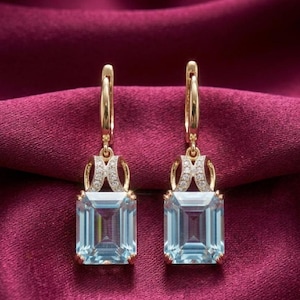 Può includere: Un paio di orecchini pendenti in oro con pietre preziose rettangolari color azzurro taglio smeraldo. Ogni orecchino presenta una chiusura a cerchio incernierata decorata con piccole pietre pavé trasparenti che collegano la pietra.