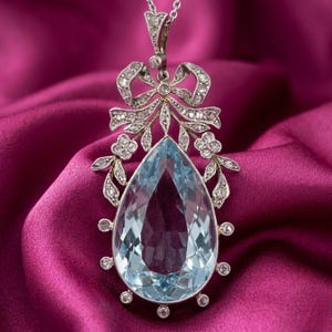 Peut inclure: Un collier pendentif en argent orné d'une grande pierre précieuse bleu clair en forme de poire. Le pendentif est orné d'un nœud et de motifs floraux, le tout incrusté de petits diamants étincelants. Le collier est présenté sur un tissu satiné rose foncé.
