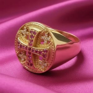 May include: A gold ring with a cross design. The ring features a large, round face with a detailed cross pattern, studded with small red and clear gemstones. The ring is set against a pink satin background, highlighting its polished gold finish.