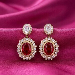 Puede incluir: Un par de pendientes dorados con una piedra central ovalada de color rojo rubí, rodeada por un halo de pequeños cristales transparentes. Los pendientes tienen un cristal más pequeño y transparente en la parte superior, sobre un fondo rosa.