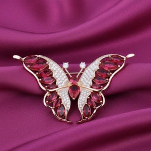 May include: A gold-toned butterfly brooch with ruby-red and clear crystal accents. The brooch features a symmetrical design with ruby-red oval-shaped stones on the wings and clear crystals in the center. The brooch is set against a deep purple background.