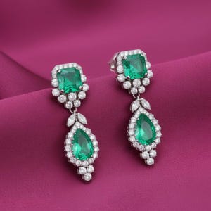 Pode incluir: Um par de brincos pendurados elegantes com pedras preciosas verdes esmeralda e um halo de pequenas pedras brancas. Os brincos têm uma parte superior quadrada e uma gota em forma de pêra, tudo em metal prateado.