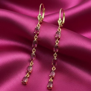 May include: A pair of gold drop earrings featuring oval, deep red gemstones. The earrings hang vertically, with each gemstone connected by small gold links. The background is a rich, pink satin fabric.