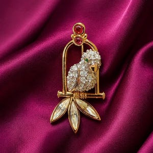 May include: A gold-toned parrot brooch with a cage design. The parrot is encrusted with clear crystals and has a green eye. Two red gemstones are at the top of the cage, and three clear crystal leaves are at the bottom.