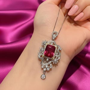 Puede incluir: Un collar de plata con un colgante grande y rectangular de piedra preciosa rojo rubí. El colgante está rodeado de piedras preciosas más pequeñas y transparentes y de intrincados filigranas de plata. Una piedra preciosa redonda más pequeña cuelga debajo.