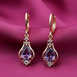 Puede incluir: Un par de pendientes colgantes dorados con una piedra preciosa púrpura en forma de lágrima y pequeñas piedras transparentes. Los pendientes tienen un cierre de palanca y se muestran sobre un fondo de satén rosa.