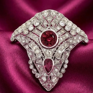 Può includere: Un'ornata spilla vintage con un design a forma di ventaglio. La spilla presenta una grande gemma rotonda di colore rosso intenso al centro, con una gemma rossa più piccola a forma di goccia sottostante. La spilla è incastonata con numerose piccole gemme trasparenti.