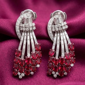 May include: A pair of silver-toned earrings featuring a fan-like arrangement of baguette-cut clear stones and multiple oval-cut deep red stones. The upper section is adorned with small round clear stones, creating a cascading, multi-layered design.