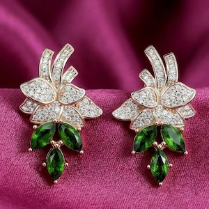 May include: A pair of gold-toned floral earrings. The earrings feature a flower design with clear, sparkling stones and three green, teardrop-shaped gemstones. The earrings are set against a pink background.