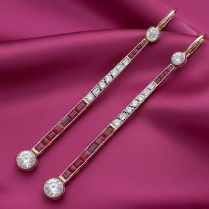 May include: A pair of gold earrings with a linear design. Each earring features a row of square red stones, alternating with clear, rectangular and round stones. The earrings have a hook closure and a large round stone at the bottom and top.