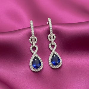 Peut inclure: Boucles d'oreilles pendantes élégantes avec des pierres précieuses bleues profondes en forme de poire, entourées d'un halo de pierres blanches scintillantes. Les boucles d'oreilles ont un design délicat et lié avec une fermeture à tige. Le bijou est posé sur un fond fuchsia vif.