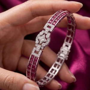 May include: A silver-toned bracelet featuring a series of square-cut deep red rubies arranged in two parallel rows. The center of the bracelet showcases a decorative circular link set with small clear white diamonds in a floral-like pattern.