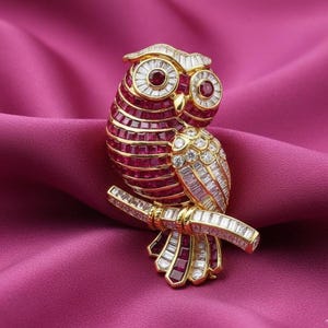 May include: A gold-toned owl brooch with ruby and diamond accents. The owl is perched on a branch, with ruby-colored stones forming the body and diamond-like stones outlining the eyes and wings. The brooch is set against a pink background.