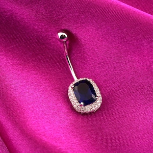 May include: A silver belly button ring featuring a dark blue, rectangular gemstone, encircled by small, clear stones. The ring is set against a vibrant, fuchsia-coloured fabric.