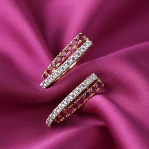 Peut inclure: Une paire de boucles d'oreilles dorées avec un design à double bande. Chaque bande est ornée d'une rangée de petites pierres précieuses rouges rondes et d'une rangée de pierres claires et scintillantes. Les boucles d'oreilles reposent sur un tissu rose foncé et soyeux.