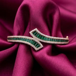 Może przedstawiać: Otwierana bransoletka typu bangle z rzędem szmaragdowozielonych kamieni o szlifie bagietkowym, otoczonych małymi, przezroczystymi kamieniami w oprawie pavé. Metalowa rama jest w kolorze złota i ma zachodzące na siebie, asymetryczne otwarcie.