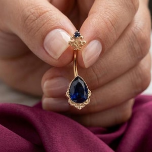 May include: A gold navel ring featuring a pear-shaped dark blue gemstone at the base and a smaller round blue gemstone at the top. The setting includes intricate filigree metalwork surrounding both stones, connected by a curved gold barbell shaft.