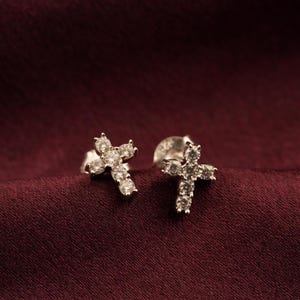 May include: A pair of silver-colored cross stud earrings. Each earring features a cross design encrusted with small, clear gemstones. The earrings are displayed on a deep red fabric background.