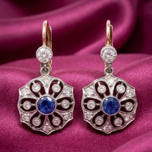 以下が含まれることがあります： ゴールドとシルバーのデザインのヴィンテージスタイルのイヤリングのペア。各イヤリングは、小さなクリアストーンに囲まれた中央の青い宝石を特徴としています。イヤリングは花のような模様とレバーバッククロージャーを備えています。