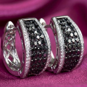 May include: A pair of silver hoop earrings with a row of black gemstones in the center, bordered by a row of smaller white gemstones. The earrings are set against a purple background.