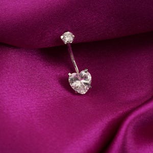 Op de afbeelding: Een zilveren navelpiercing met een hartvormige heldere edelsteen aan de onderkant en een ronde heldere edelsteen aan de bovenkant. De sieraden zijn geplaatst tegen een dieppaarse satijnen achtergrond.